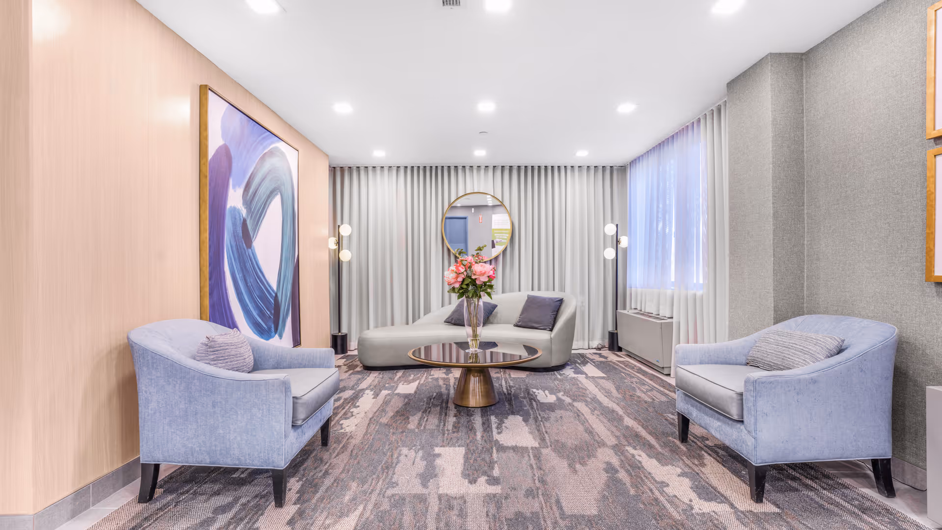 A modern and cozy living room area with two light blue armchairs facing a round coffee table with a vase of pink flowers. Behind the table is a light gray chaise lounge with two dark gray pillows. The room features a large abstract painting on the left wall, a round mirror on the back wall with gray curtains, and soft lighting from floor lamps on either side of the chaise lounge. The floor is covered with a patterned carpet in shades of gray and beige.
