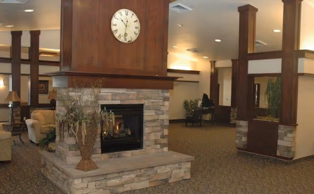 Spacious assisted-living common area with a stone fireplace and clock on a wooden mantle, seating, and a piano in the background.