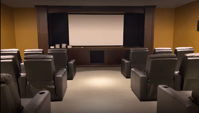 A small theater room with multiple rows of black leather recliner chairs facing a large white projection screen mounted on a dark wooden wall.