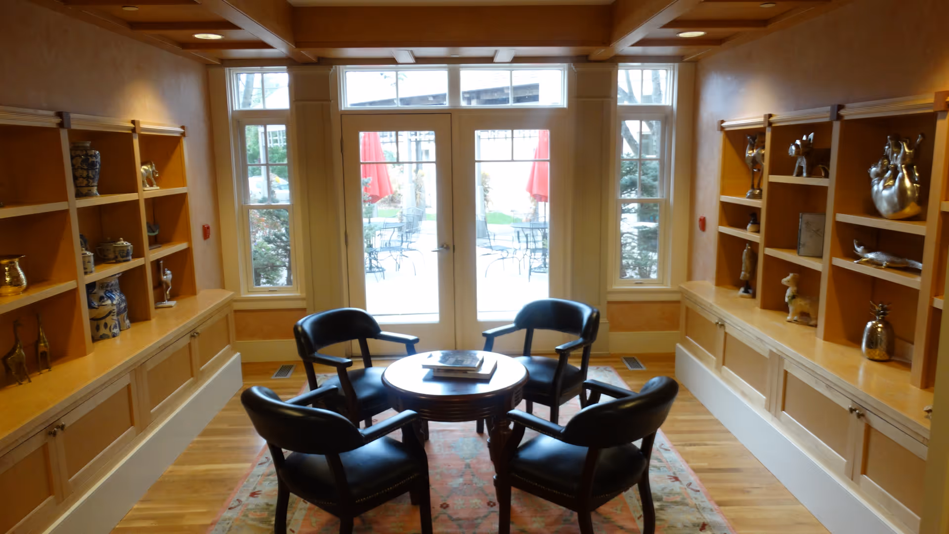 A cozy sitting area with four black leather chairs arranged around a small round wooden table on a patterned rug. The room has wooden built-in shelves on both sides displaying decorative items and vases. Large windows and glass doors at the far end provide a view of an outdoor patio with tables and red umbrellas.
