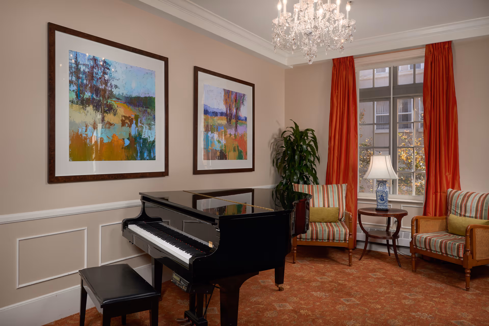 A cozy interior room featuring a black grand piano with a matching bench, two striped armchairs with green cushions, a small round wooden table with a blue and white lamp, two colorful abstract paintings on the beige wall, a large window with red curtains, a green potted plant, and a crystal chandelier hanging from the ceiling.