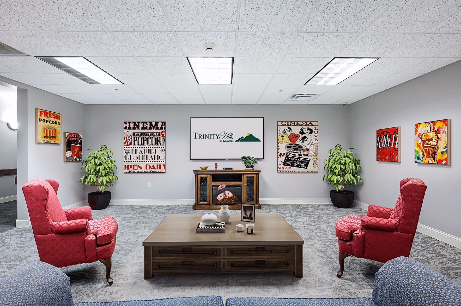 A cozy lounge area with two red upholstered armchairs facing a wooden coffee table with decorative items. The walls are decorated with colorful vintage-style movie and popcorn posters. Two large potted plants are placed in the corners. A wooden cabinet with a framed sign reading 'Trinity Hills of Knoxville' is centered on the back wall. The room has a gray carpet and a white drop ceiling with fluorescent lights.