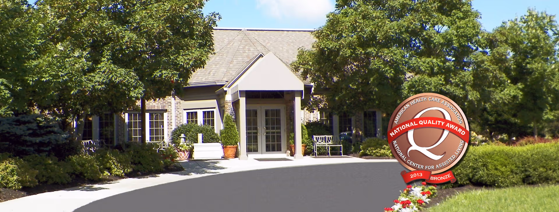 Exterior view of Burton's Ridge Assisted Living facility showing the entrance with a covered porch, surrounded by trees, bushes, and a well-maintained garden under a blue sky.