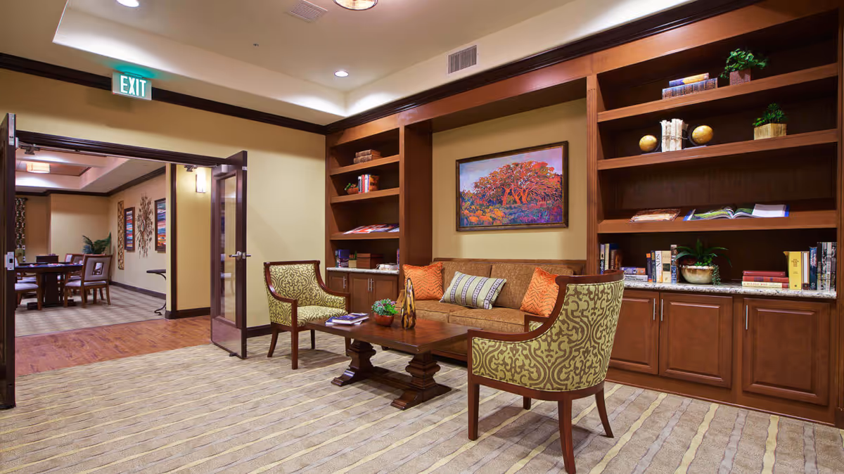 A cozy seating area with built-in wooden bookshelves, a sofa and patterned chairs around a coffee table in a senior living common room.