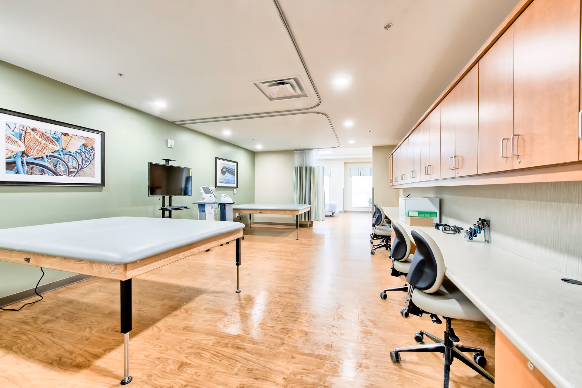 A spacious therapy or medical treatment room with two padded treatment tables, a row of chairs with a long countertop and cabinets above on the right side, a TV mounted on the green wall, and medical equipment near the back. The room has wood flooring and bright overhead lighting.