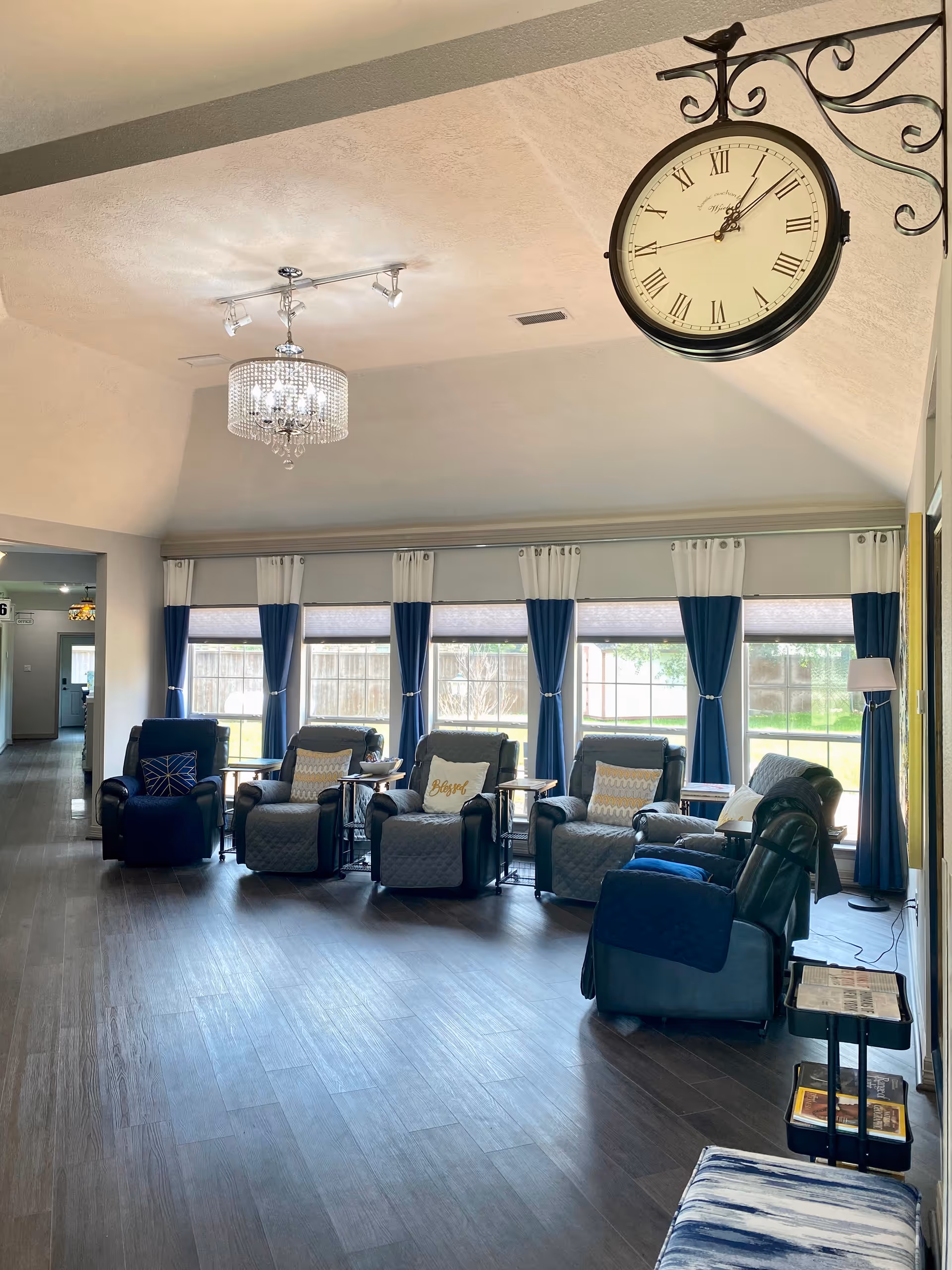 A bright and spacious living room area with five comfortable recliner chairs arranged in a row facing large windows with blue and white curtains. The room has dark wood flooring, a decorative hanging clock with Roman numerals on the wall, and a chandelier light fixture on the ceiling. There are small side tables between the chairs and a hallway visible in the background.
