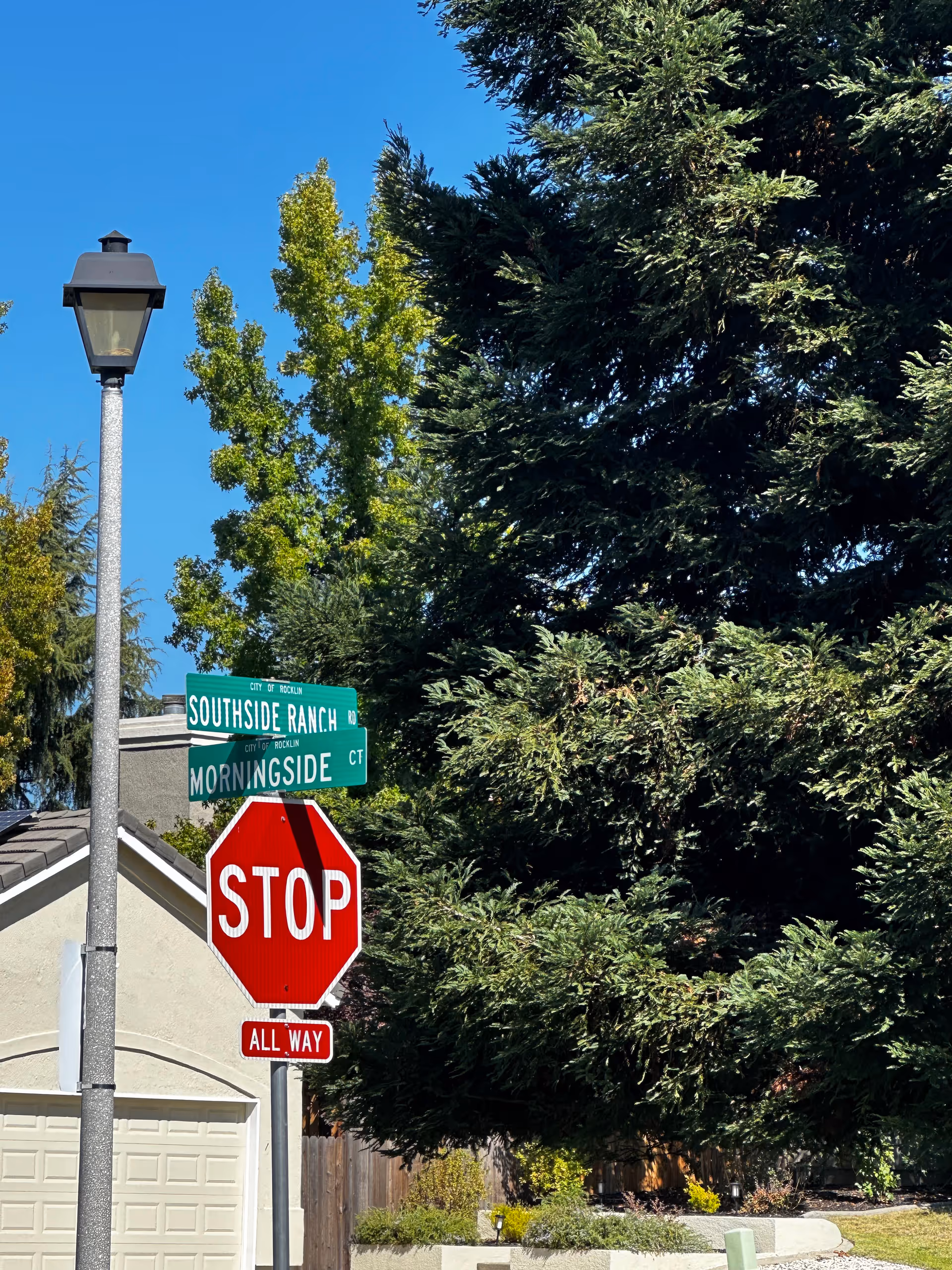 Street corner with a stop sign and street name signs for Southside Ranch Rd and Morningside Ct, surrounded by trees and a house garage in the background under a clear blue sky.