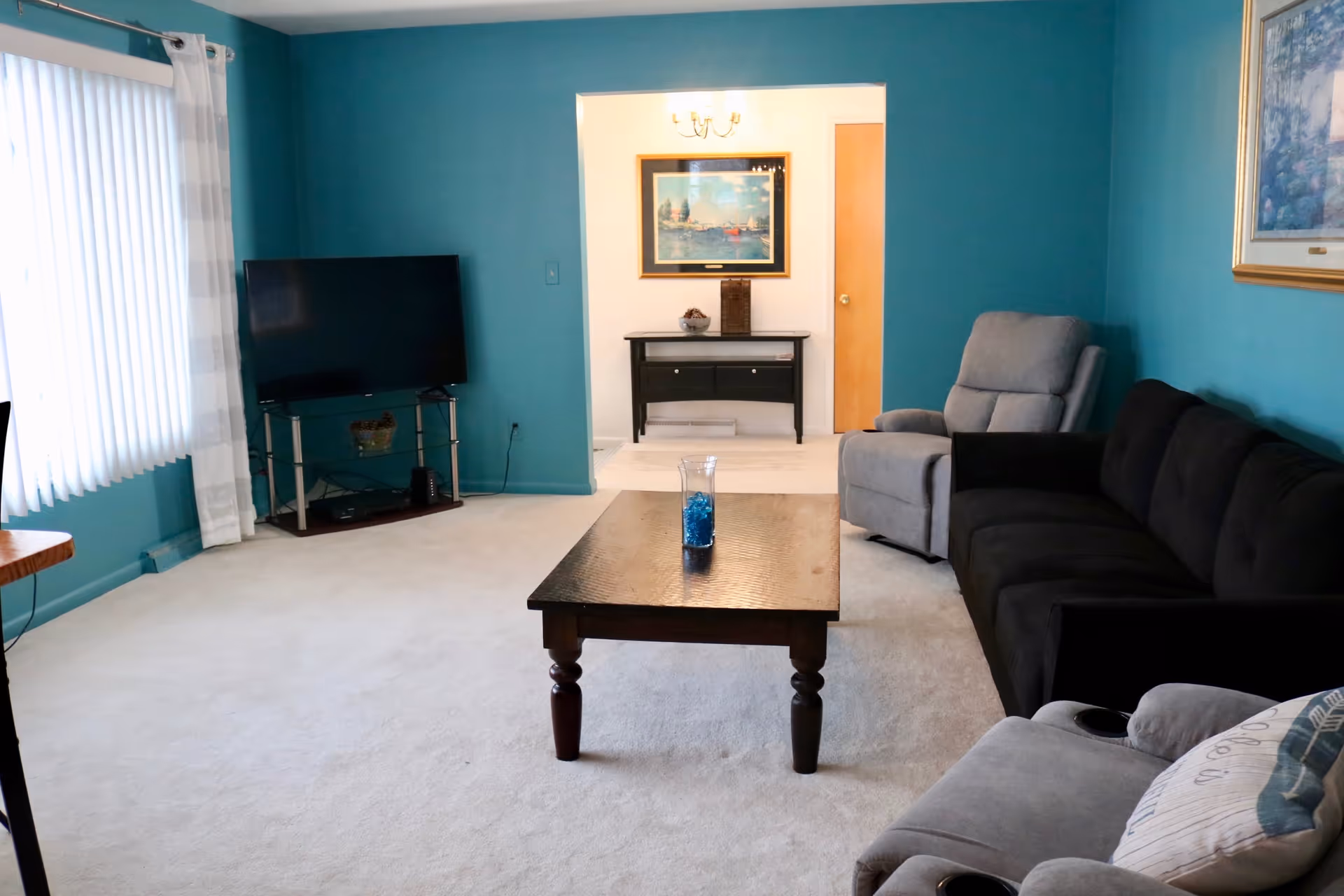 A living room with teal walls and light carpet. The room features a black sofa, two gray recliners, a wooden coffee table with a glass vase containing blue decorative stones, and a flat-screen TV on a stand near a window with vertical blinds and white curtains. In the background, there is a small black console table with decorative items and a framed painting above it.