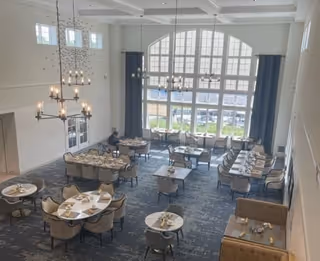 A spacious dining room with multiple tables and chairs arranged neatly. The room features large arched windows allowing natural light to fill the space, modern chandeliers hanging from a high ceiling, and a carpeted floor. Some tables are set with plates and napkins, and a person is seated at one of the tables near the windows.