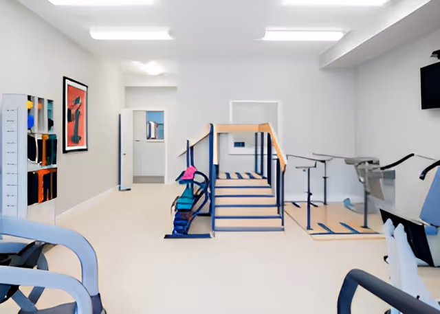 A physical therapy or rehabilitation room with exercise equipment including parallel bars, a small staircase with handrails, and various exercise machines. The room has white walls, bright overhead lighting, and a framed artwork on the left wall.