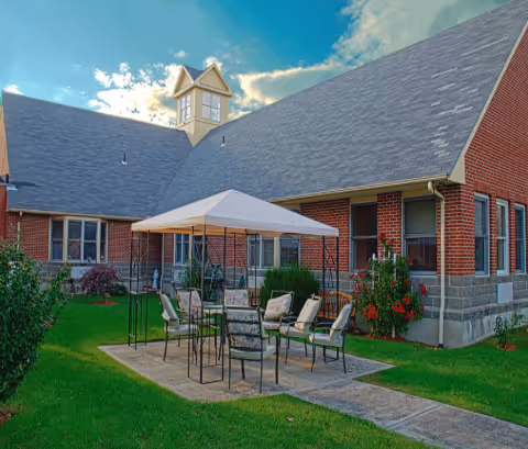 Outdoor patio area with a white canopy tent covering several chairs and tables on a paved surface, surrounded by green grass and landscaping. The background shows a brick building with multiple windows and a steep roof under a partly cloudy sky.