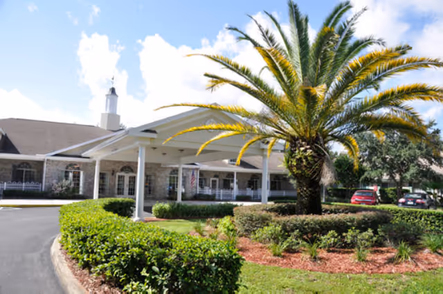 Exterior view of a senior living facility with a covered entrance, a large palm tree, well-maintained bushes, and a driveway with parked cars under a partly cloudy sky.