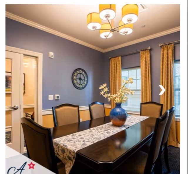 A dining room with a dark wooden table and six upholstered chairs. The table has a floral runner and a blue vase with white flowers. The walls are painted blue with a white chair rail and yellow lower section. There is a large clock on the wall and two windows with yellow curtains. A modern chandelier with yellow lampshades hangs from the ceiling.