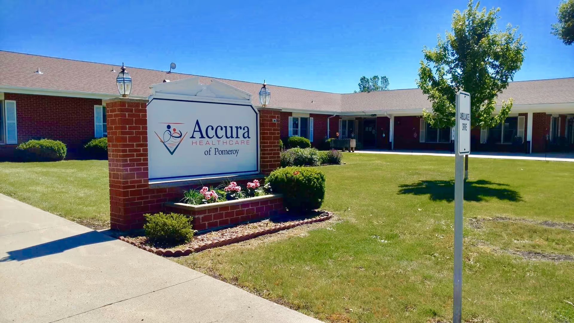 Exterior view of Accura HealthCare of Pomeroy building with a brick sign displaying the facility name. The building is single-story with a brown roof and red brick walls. There is a well-maintained lawn with bushes, flowers, and a small tree near a signpost that reads 'AMBULANCE PARKING'. The sky is clear and blue.