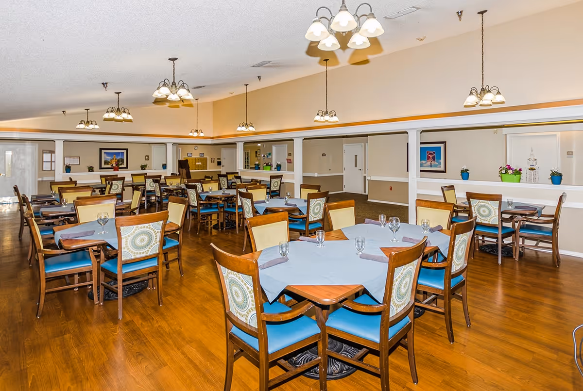 A spacious dining room with multiple wooden tables and chairs arranged neatly. Each table is covered with a light blue tablecloth and set with glasses and napkins. The room has warm wooden flooring, beige walls, and several hanging light fixtures providing ample lighting. There are framed pictures and potted plants on the walls and ledges, creating a welcoming atmosphere.