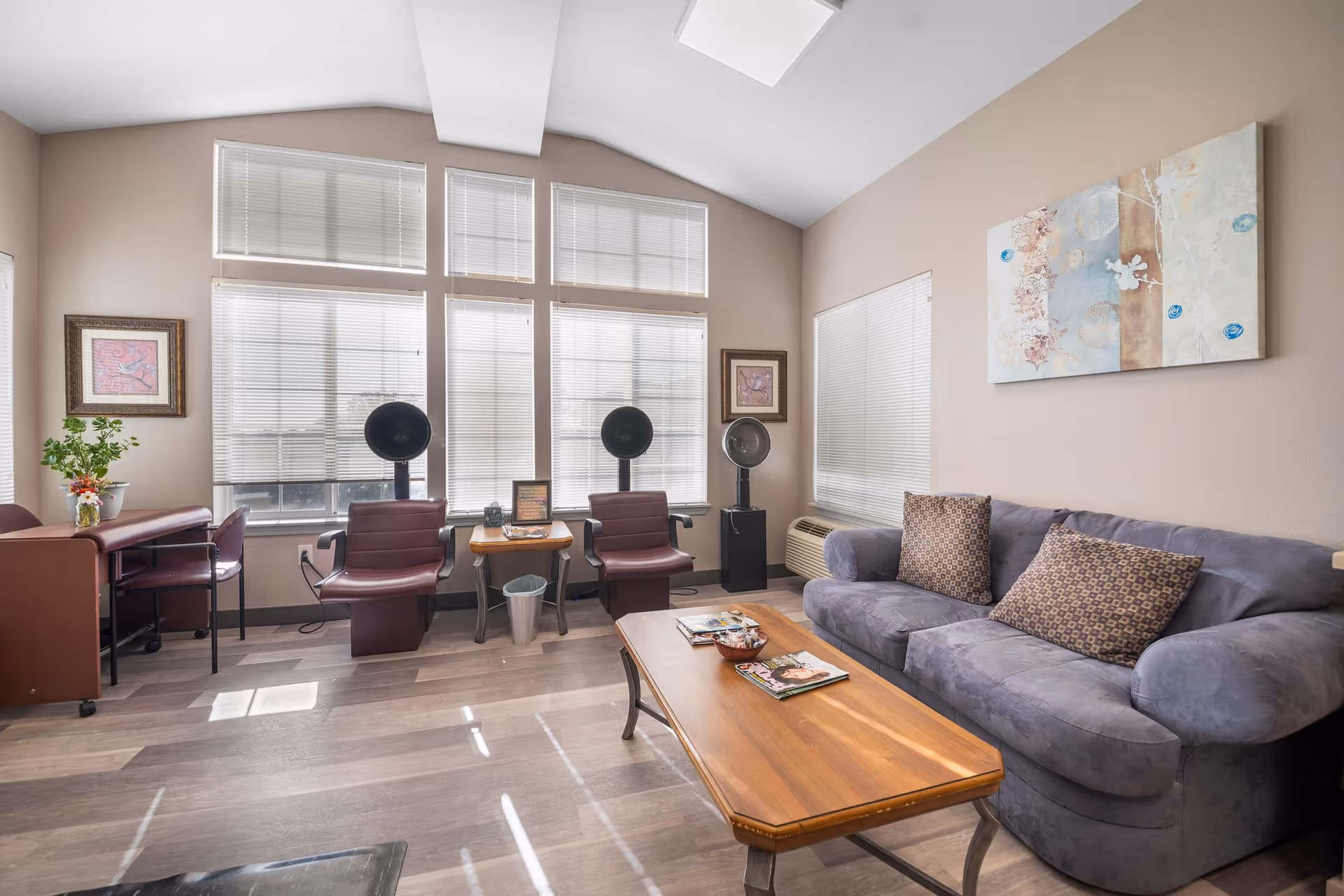 Sunlit communal lounge with a blue sofa, coffee table, two salon chairs with hooded hairdryers, and large windows.