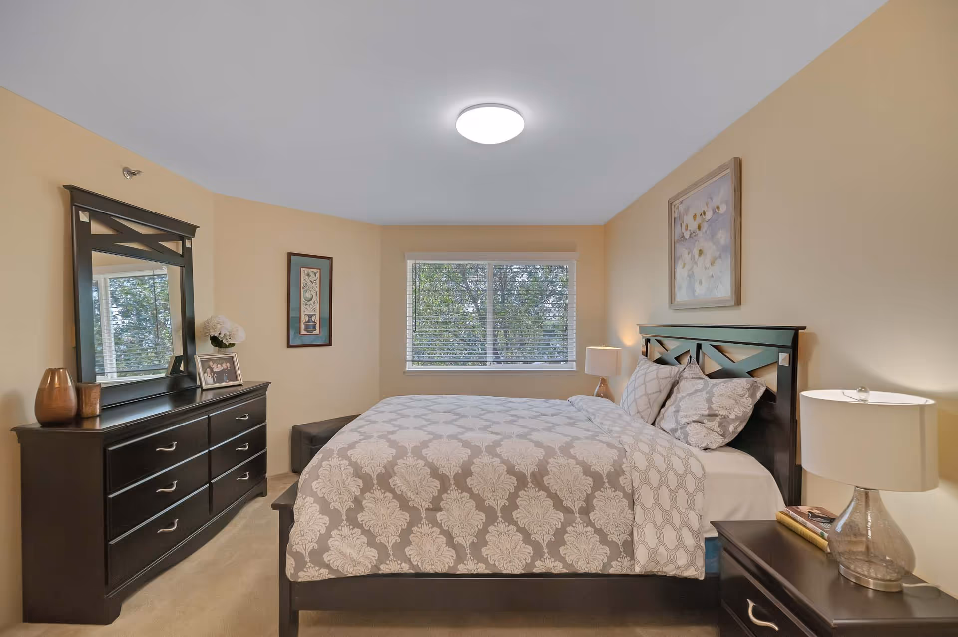 A neatly arranged bedroom with a large bed covered in a patterned beige and white comforter. The room features a dark wooden headboard, matching dresser with a mirror, and a nightstand with a lamp and books. A window with white blinds lets in natural light, and there are framed artworks on the walls.