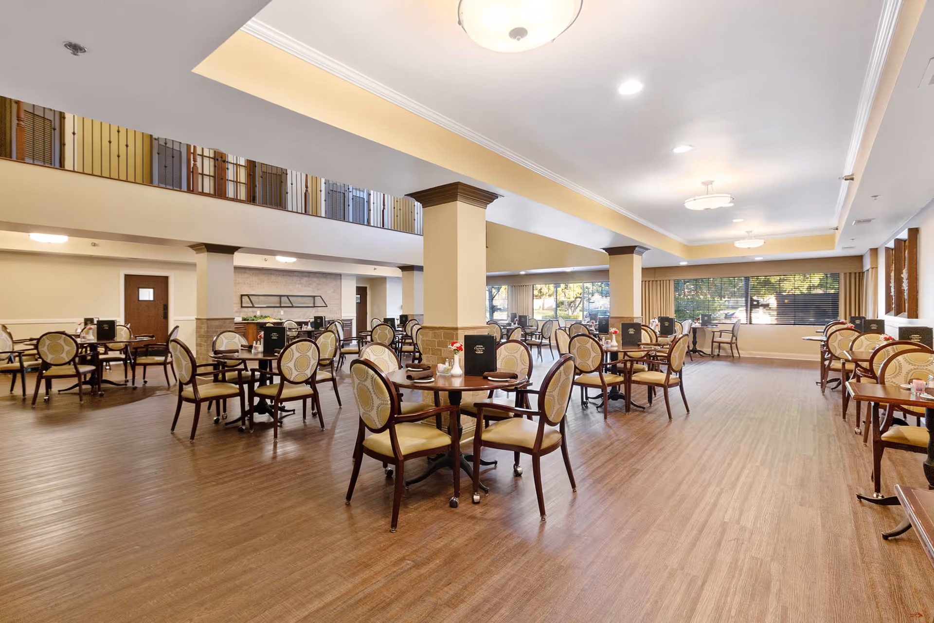 Spacious dining room with multiple round tables and chairs, wood flooring, and large windows letting in natural light.