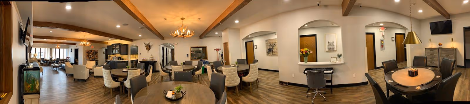 Panoramic view of a spacious senior living common area with multiple dining tables and chairs, lounge seating, exposed wooden beams, decorative lighting, a piano, and an aquarium.