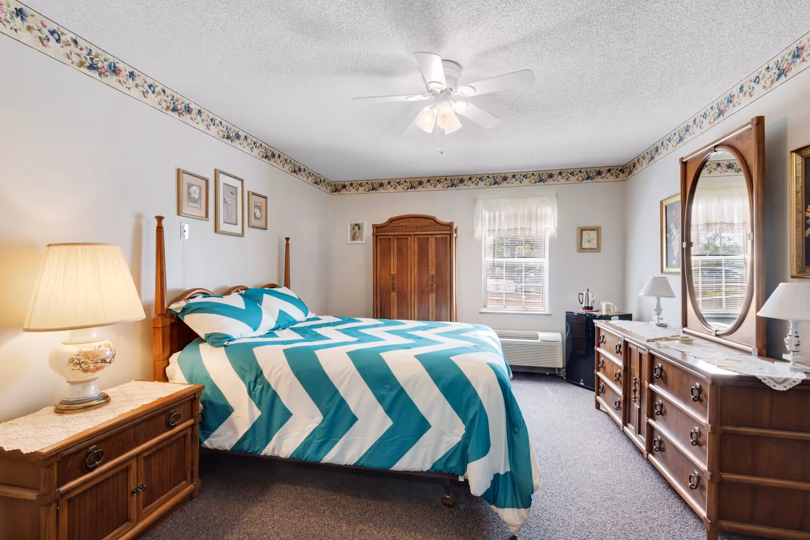A cozy bedroom in Grandview Retirement Center featuring a bed with a teal and white chevron patterned comforter, wooden nightstand with a floral lamp, wooden dresser with an oval mirror, a wooden armoire, and a window with sheer curtains letting in natural light. The room has floral wallpaper border near the ceiling and carpeted floor.