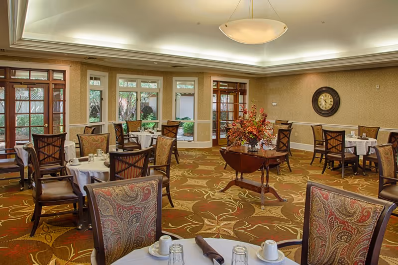 A spacious dining room in a senior living facility with multiple round tables covered with white tablecloths, set with cups, glasses, and napkins. The room features patterned carpet, upholstered chairs with intricate designs, large windows, and glass doors letting in natural light. A decorative table with a floral arrangement is placed in the center, and a large wall clock is visible on the far wall.
