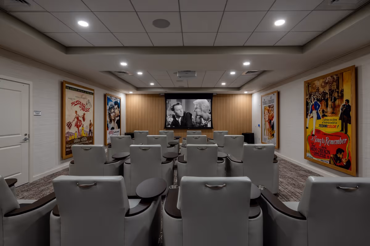 A small movie theater room with multiple rows of comfortable white leather recliner chairs facing a large screen showing a black and white movie. The walls are decorated with framed vintage movie posters including The Sound of Music, Grease, and A Song to Remember. The ceiling has recessed lighting and a projector mounted in the center.
