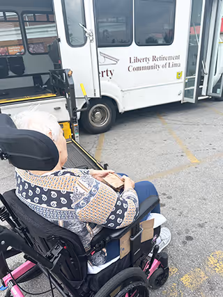 An elderly person in a wheelchair sits in a parking lot next to a Liberty Retirement Community of Lima shuttle bus.