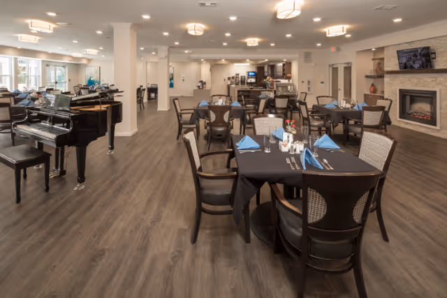 A spacious dining room with multiple tables covered in black tablecloths and set with blue napkins, silverware, and glasses. There is a grand piano on the left side and a fireplace with a TV mounted above it on the right. The room has wood flooring and is well-lit with ceiling lights.