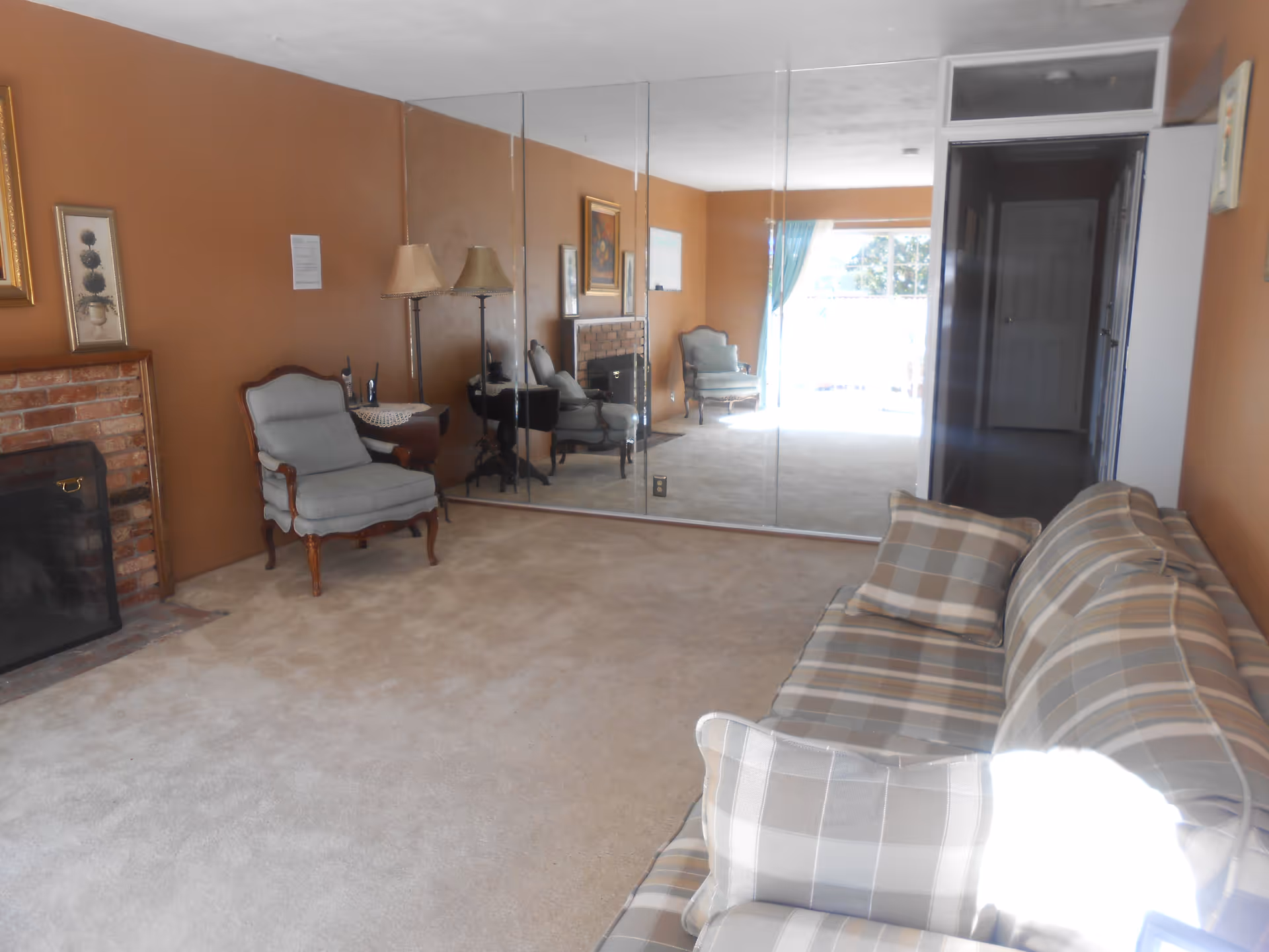 A living room with beige carpet and brown walls. There is a plaid sofa with matching pillows on the right side, a single upholstered armchair near a brick fireplace on the left, and a floor lamp next to the chair. The far wall is covered with large mirrors reflecting the room. A window with blue curtains lets in natural light at the back, and there is an open doorway leading to a hallway.