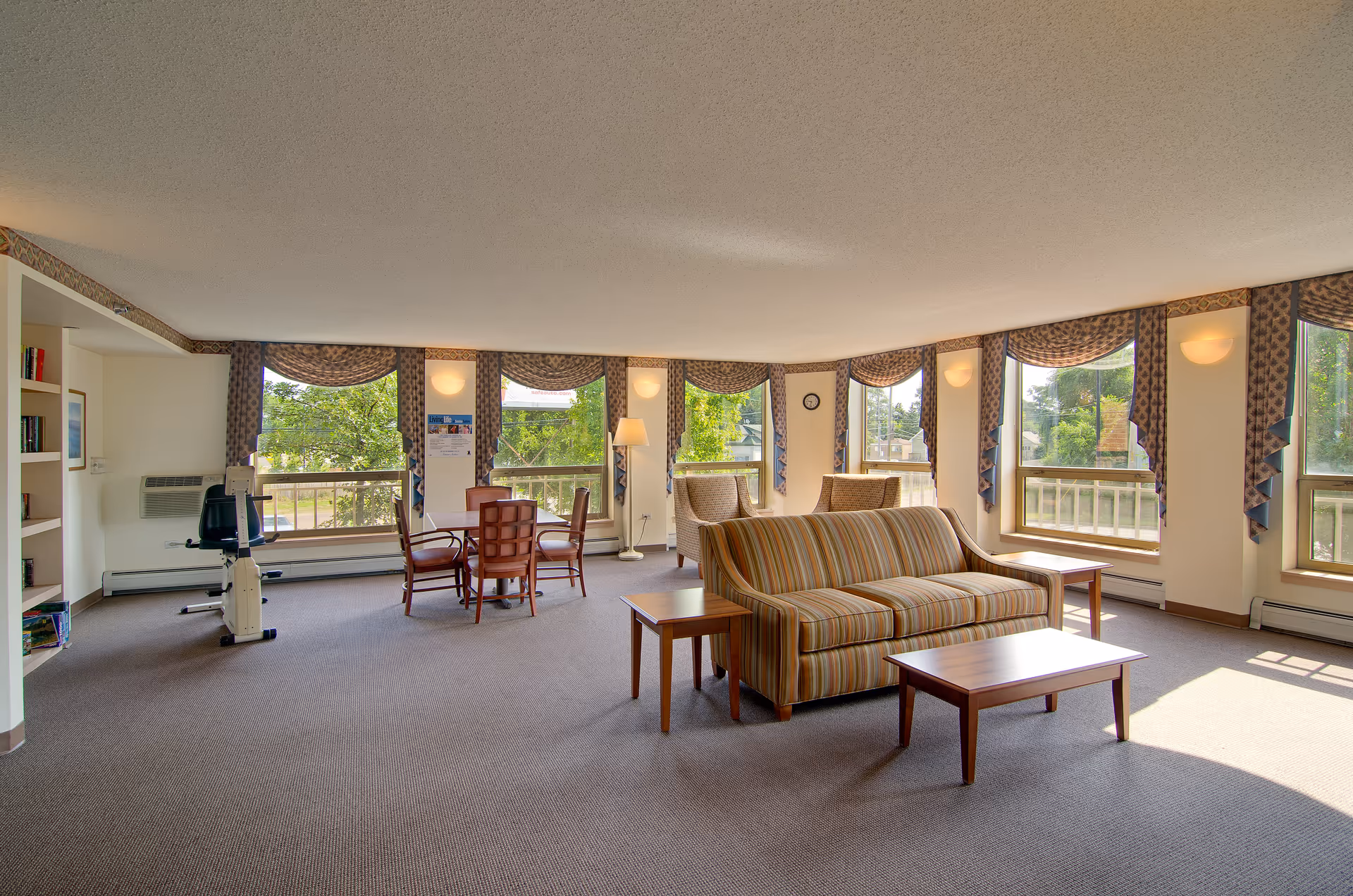 A spacious, well-lit living room area with large windows adorned with patterned valances allowing natural light to fill the room. The room features a striped sofa with two matching armchairs, a wooden coffee table, and side tables. There is a small dining table with four chairs near the windows, a floor lamp, and an exercise bike near a built-in bookshelf.