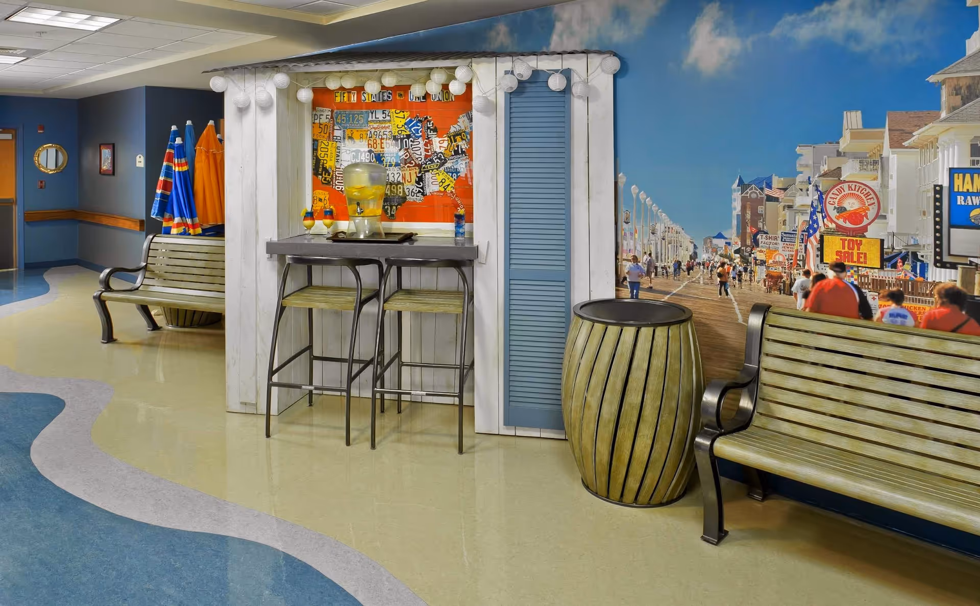 Indoor seating area with two wooden benches and a small bar table with two stools. The wall behind features a colorful mural of a busy boardwalk scene with people walking and various storefronts. There is a decorative barrel next to one bench and a water dispenser on the bar table. The floor has a wavy blue and beige pattern.
