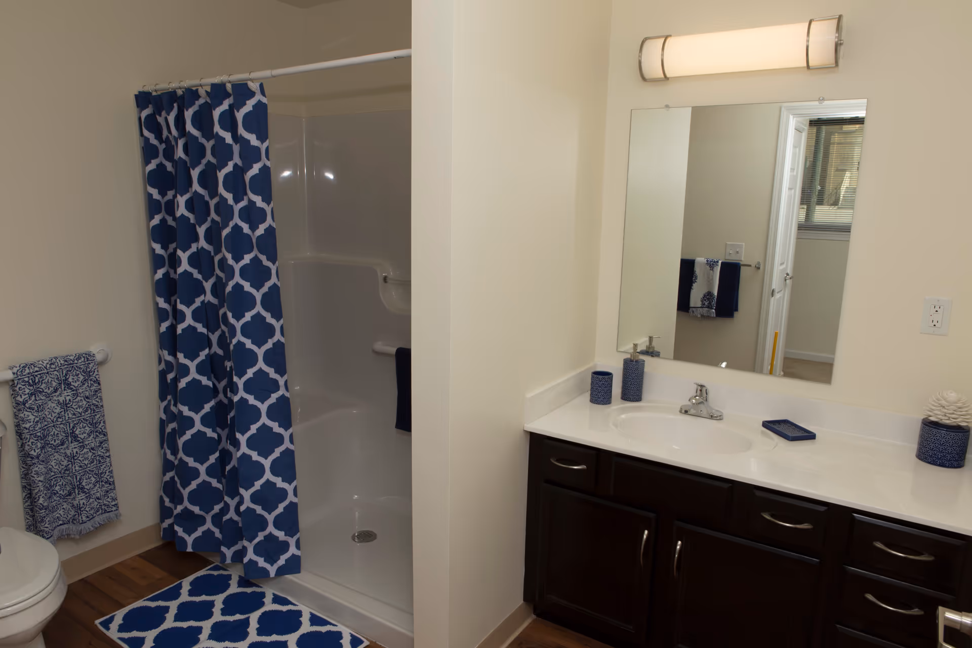 A bathroom with a white shower stall partially covered by a blue and white patterned shower curtain. There is a matching blue and white rug on the wooden floor in front of the shower. To the right is a white countertop with a sink, dark wood cabinets underneath, and a large mirror above. On the countertop are blue decorative items including a soap dispenser, a cup, and a small tray. A towel rack with a blue and white patterned towel is visible next to the toilet on the left side.