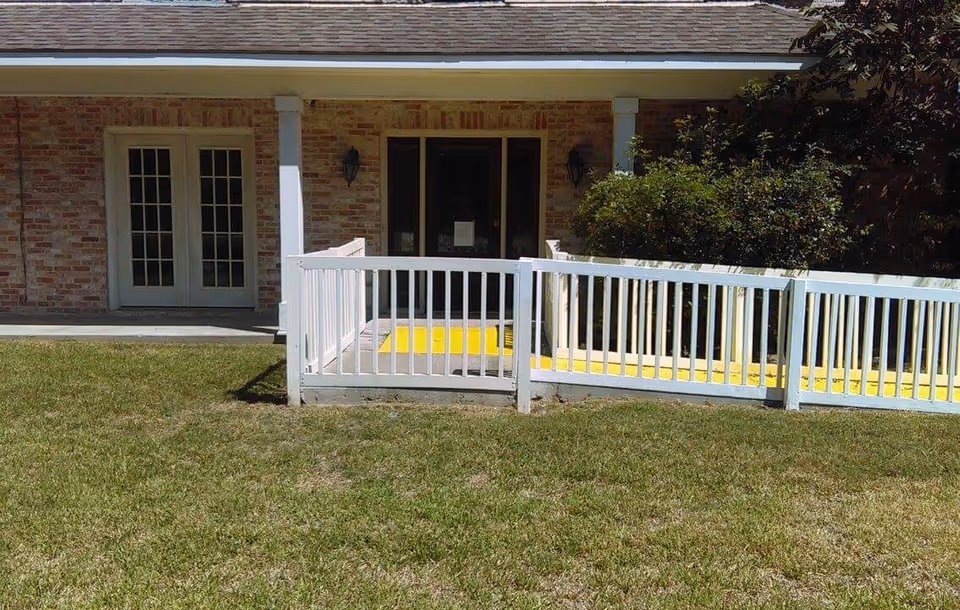 Exterior view of a brick building with a white railing and a yellow ramp leading to a door. There is a grassy lawn in the foreground and some bushes near the entrance.