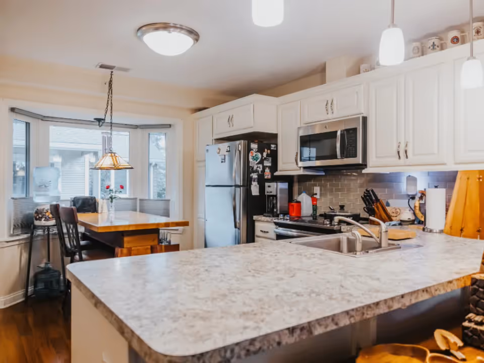 Bright kitchen with white cabinets, stainless steel refrigerator and microwave, a large marble countertop with a sink, and a wooden dining table with chairs near a bay window. A hanging light fixture is above the dining table, and various kitchen items are on the counters.