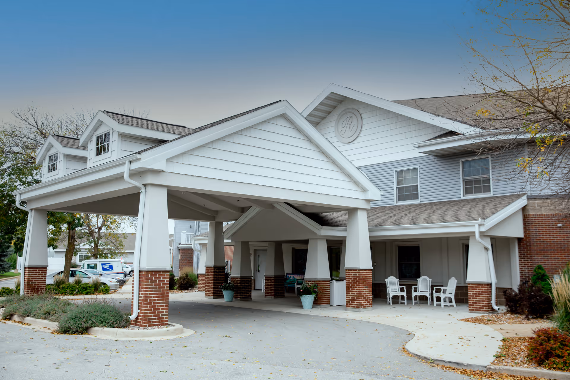 Covered porte-cochère entrance of a multi-story senior living building with brick columns, outdoor seating, and surrounding landscaping.