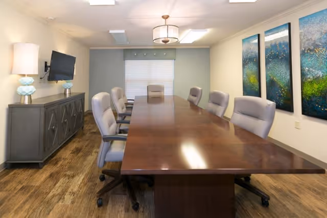 A conference room with a long wooden table surrounded by eight gray office chairs on wheels. The room has wood flooring, a sideboard with two decorative lamps, a wall-mounted TV, and three abstract paintings on the wall. A window with blinds and a valance is at the far end of the room.