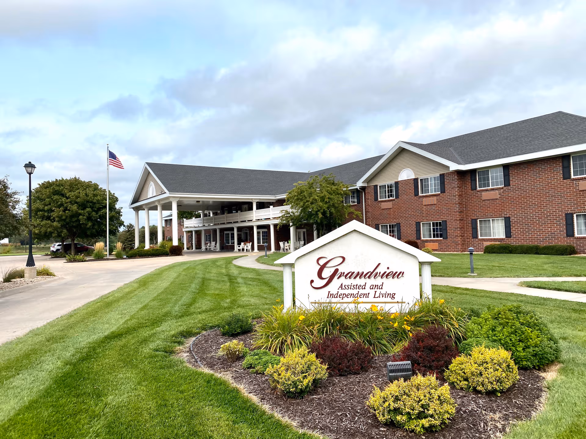 Exterior view of Grandview Assisted and Independent Living facility showing a large brick building with white columns at the entrance, a well-maintained lawn, landscaped bushes and flowers, an American flag on a flagpole, and a sign in the foreground with the facility's name.