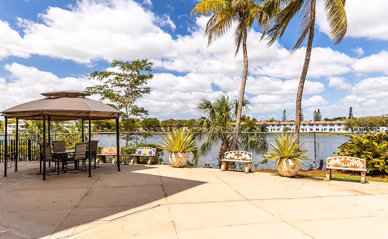 Lakeside outdoor patio with a gazebo, seating benches, potted plants, and palm trees under a partly cloudy sky.