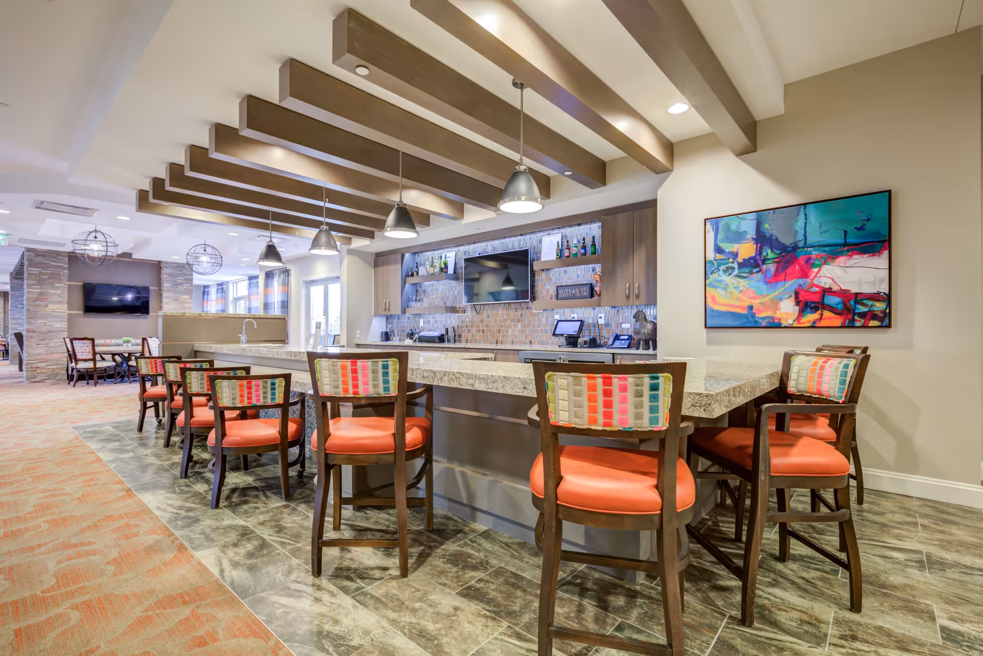 Interior view of a senior living facility lounge area featuring a long granite countertop with several high chairs upholstered in orange and multicolored fabric backs. Above the counter are pendant lights and wooden ceiling beams. Behind the counter is a bar area with shelves holding bottles and a mounted flat-screen TV. The floor is a mix of tile and carpet, and there is colorful abstract artwork on the wall.