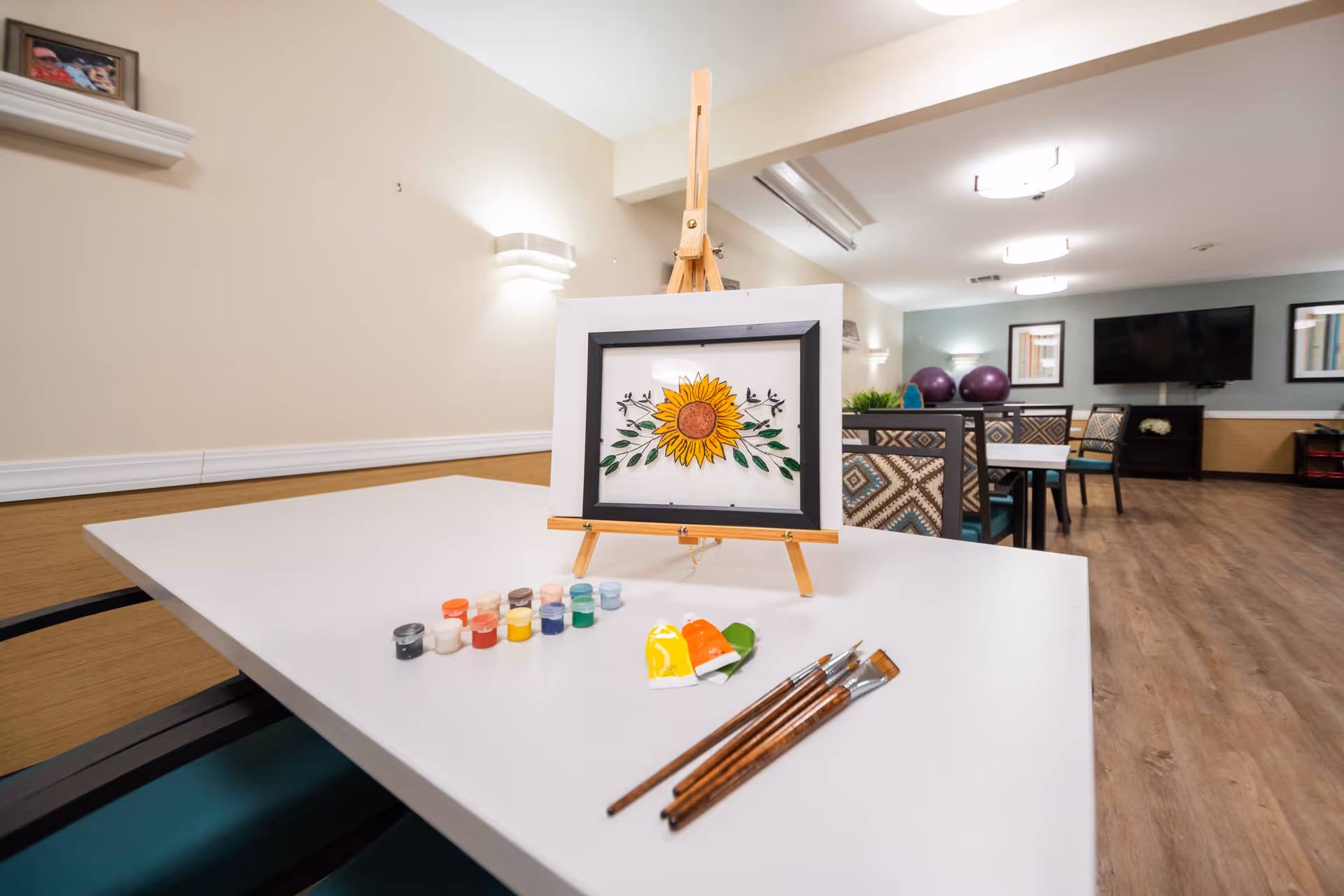 A bright room with a white table displaying a framed sunflower painting on an easel, surrounded by small paint pots, paint tubes, and brushes. In the background, there are chairs with patterned upholstery, exercise balls, a large TV mounted on the wall, and framed pictures.