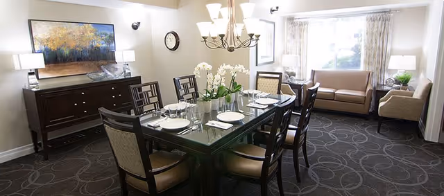 A well-lit dining room with a rectangular glass-top table set for six people with plates, glasses, and napkins. The room features a chandelier above the table, a sideboard with two lamps and a large framed painting on the wall, a clock, and a seating area with a sofa and an armchair near a window with curtains.