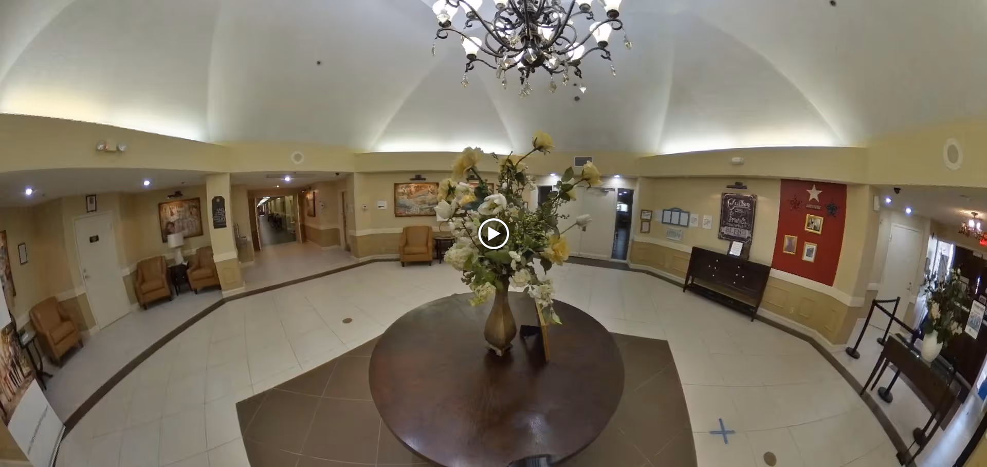 A spacious lobby area with a round wooden table in the center holding a vase of flowers. The room has beige walls with white trim, several armchairs along the walls, framed artwork, and a chandelier hanging from the ceiling. There are hallways and doors leading to other parts of the facility.