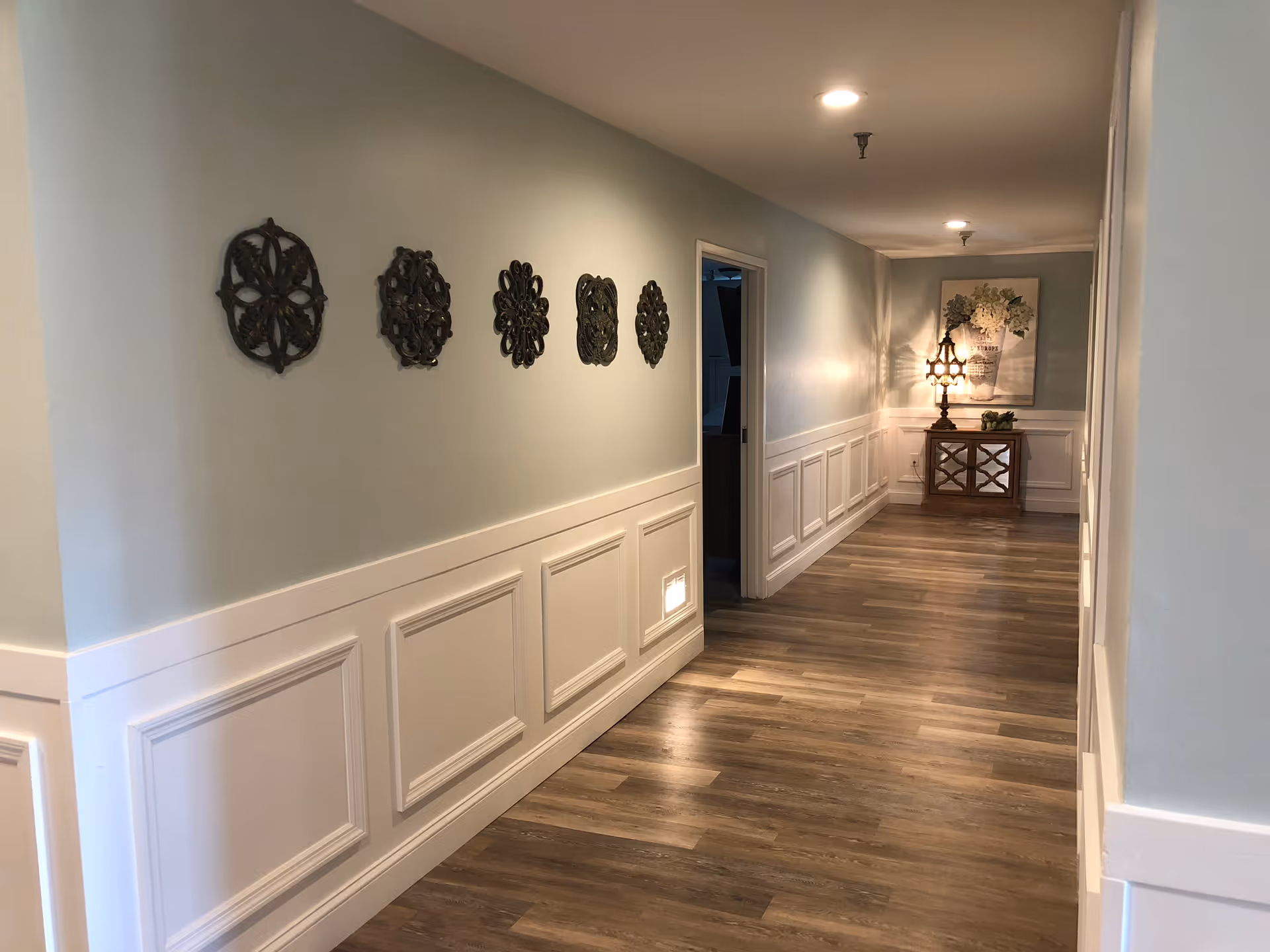 A well-lit hallway with light blue walls and white wainscoting. The floor is wood with a natural finish. Five decorative metal wall hangings are arranged in a row on the left wall. At the end of the hallway, there is a small wooden cabinet with a decorative lamp and a floral painting above it. Two doorways are visible on the right side of the hallway.