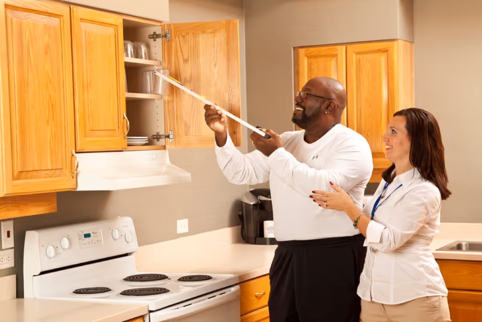 Two people in a kitchen, the man using a tape measure to check an open upper cabinet while the woman watches.