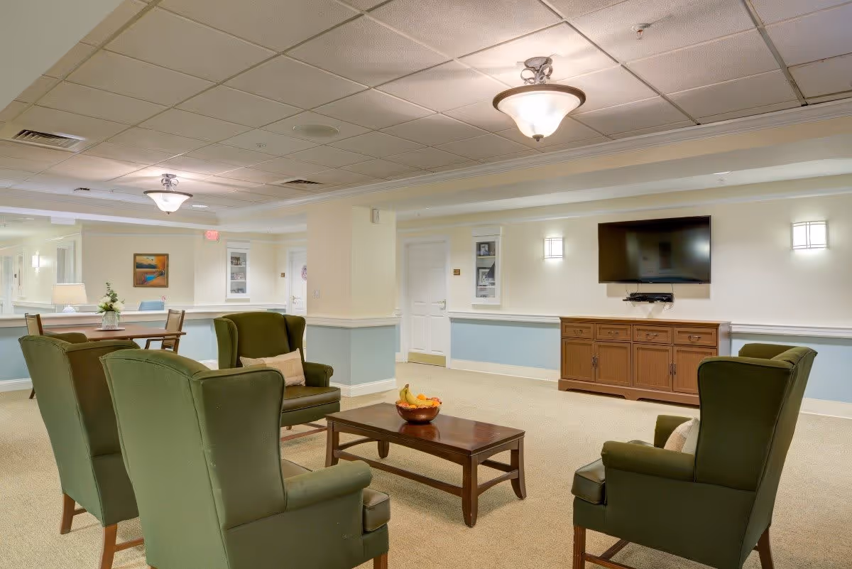 A cozy common area with four green upholstered armchairs arranged around a wooden coffee table holding a bowl of fruit. A wooden cabinet with a flat-screen TV mounted above it is against the wall. The room has soft lighting with ceiling fixtures and wall sconces, light-colored walls with a blue wainscot, and a beige carpeted floor. In the background, there is a small dining table with chairs and a lamp on a side table.