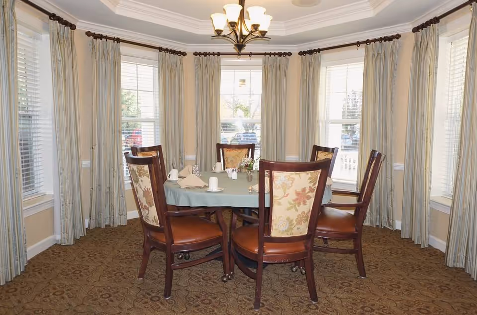 A dining room with a round table covered with a light blue tablecloth, set with cups, napkins, and a small flower arrangement. Six wooden chairs with floral upholstered backs and brown leather seats surround the table. The room has beige walls, patterned carpet, multiple windows with white blinds, and light-colored curtains. A chandelier with five lights hangs from the ceiling.