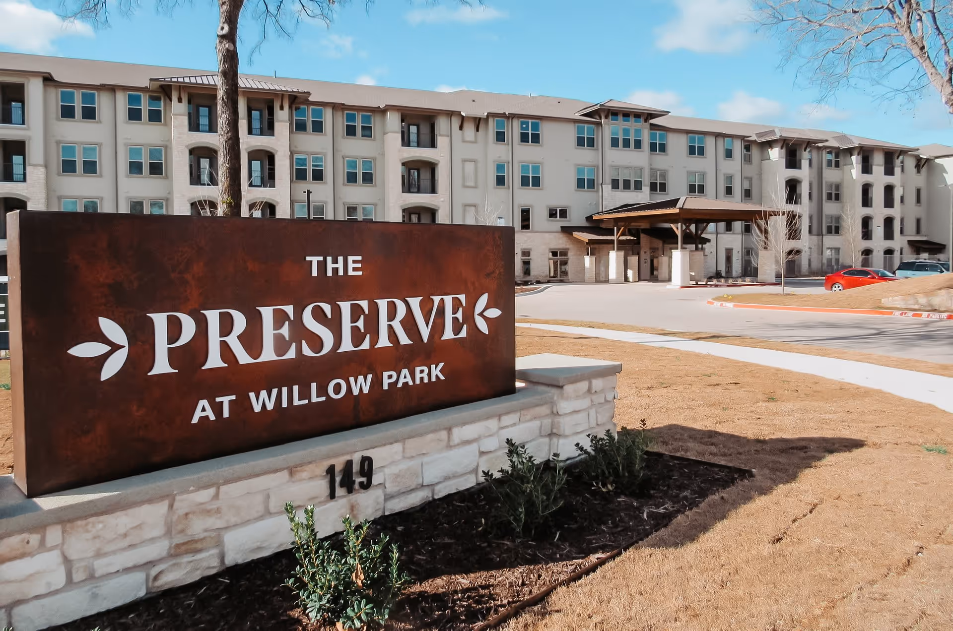 Exterior view of a multi-story senior living facility named The Preserve at Willow Park with a large sign in the foreground displaying the facility's name and address number 149. The building has multiple windows, balconies, and a covered entrance area. The sky is clear with some clouds.