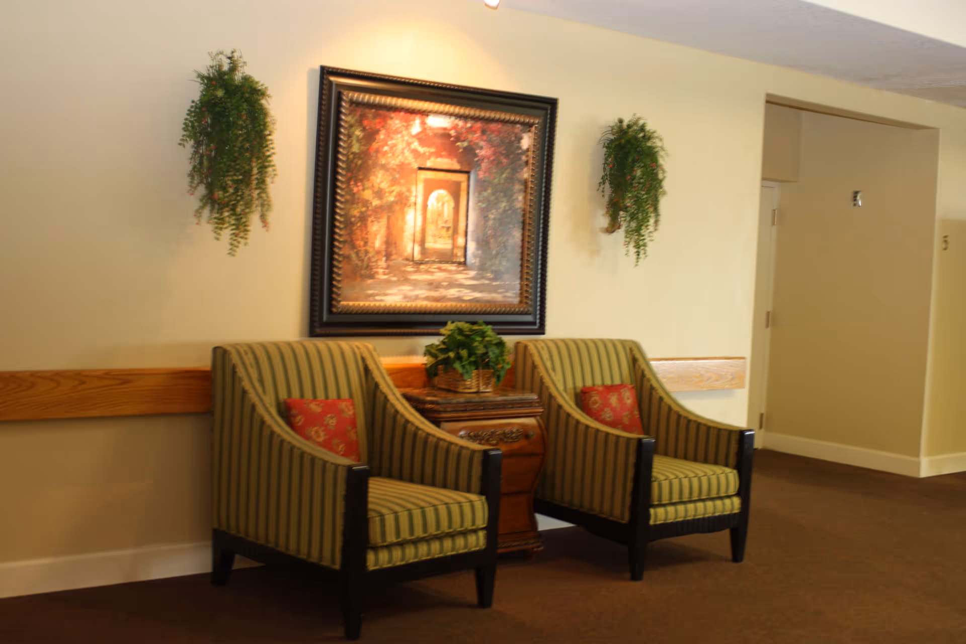Two green-striped armchairs flank a small wooden table beneath a framed painting and hanging plants in a hallway seating area.