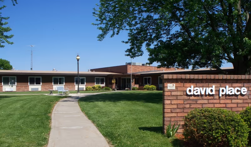 Exterior view of a single-story brick building with a sign that reads 'david place' in white letters. The building is surrounded by a well-maintained lawn, a concrete walkway, a lamppost, and several trees under a clear blue sky.