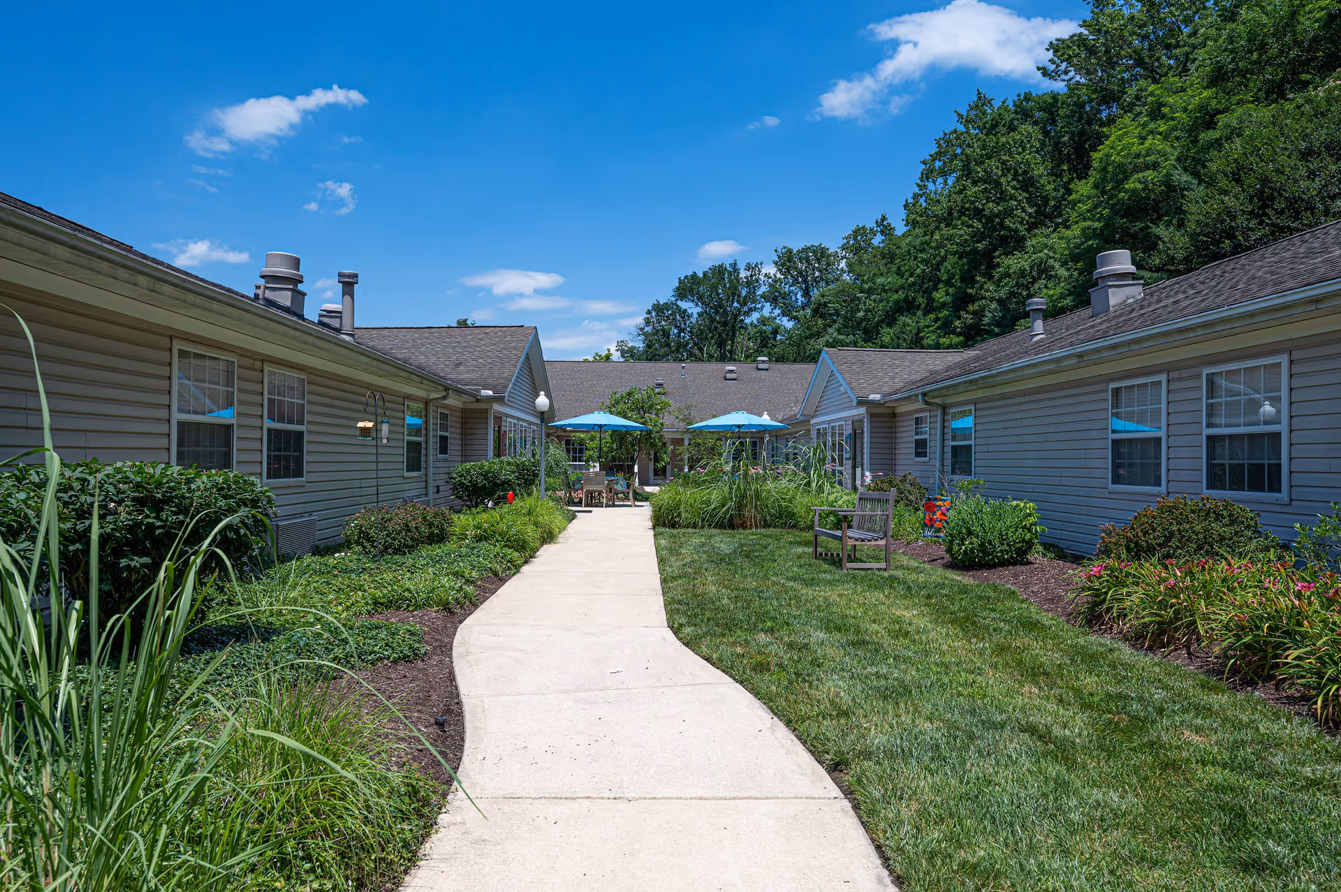 A sunny outdoor courtyard area at Arden Courts - ProMedica Memory Care Community in Towson, featuring a concrete pathway flanked by green grass, landscaped bushes, and flowers. There are two buildings on either side with beige siding and multiple windows. In the distance, there are patio tables with blue umbrellas and chairs, surrounded by trees and a clear blue sky with a few clouds.